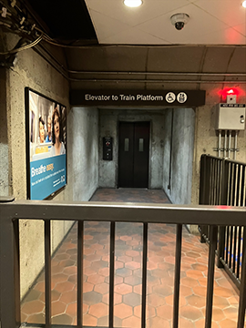 A picture of the path to an elevator. In the foreground there is an emergency exit gate. There is an advertisement on the wall to the left and a section of railing to the right. In the background is the elevator.